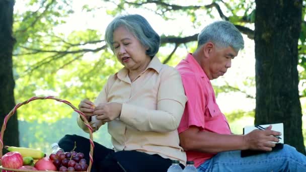 Życie na emeryturze: Starsza para jest szczęśliwa i szczęśliwa siedząc w ogrodzie pod cienistymi drzewami, robiąc zajęcia rekreacyjne, jedząc owoce i rysując zdjęcia natury. - Materiał filmowy, wideo