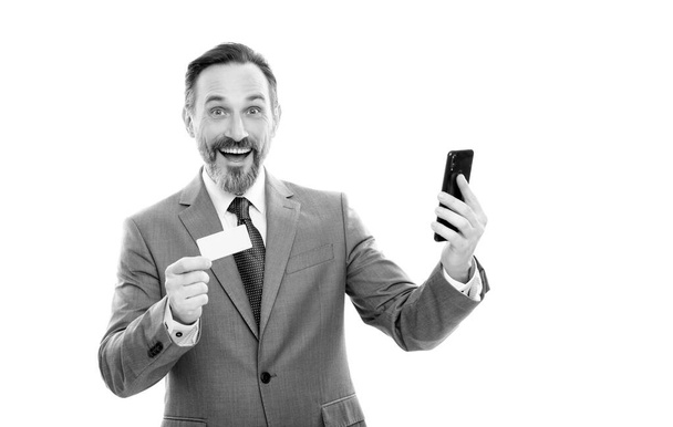 スーツの幸せな成熟したオフィスマネージャーは白に隔離されたデビットカードとスマートフォンを保持し、インターネット購入. - 写真・画像