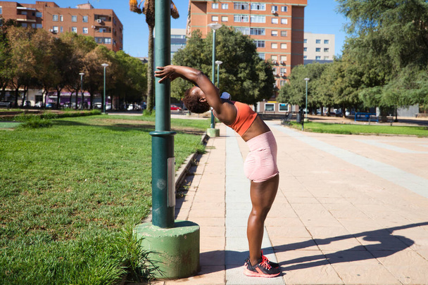 Jeune et belle Afro-Américaine au corps sculpté s'étirant dans le parc avant l'entraînement. Elle porte un haut orange et des leggings orange. Concept de sport et de santé. - Photo, image