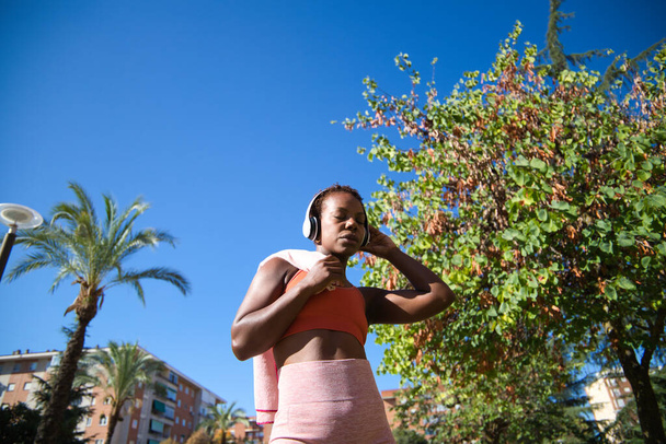 Afroameričanka, která sportuje v parku, nosí přes rameno ručník a poslouchá hudbu s bílými sluchátky. Je oblečená v oranžovém svršku. Pojetí sportu a zdraví. - Fotografie, Obrázek