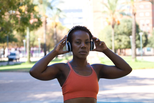 Young and beautiful Afro American woman with sculpted body sitting in the park before training. She is dressed in an orange top and tights and listens to music with white headphones. Sport and health. - Photo, Image