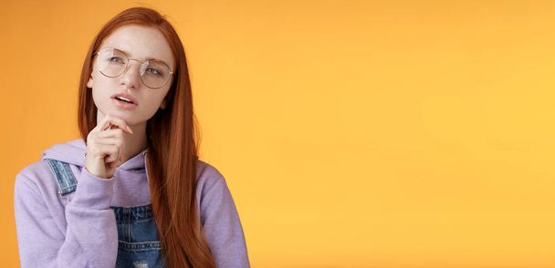 Thoughtful young creative smart redhead girl thinking figure out important thing standing upper left corner squinting thinking get clue touch chin thinking, pondering choice, orange background. - Photo, Image