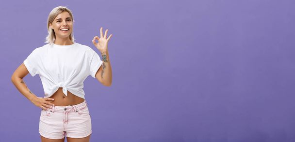 Excellent I like it. Portrait of satisfied good-looking happy girl with fair hair in white t-shirt and shorts showing ok or perfect gesture and smiling broadly holding hand on waist over purple wall. - Photo, Image