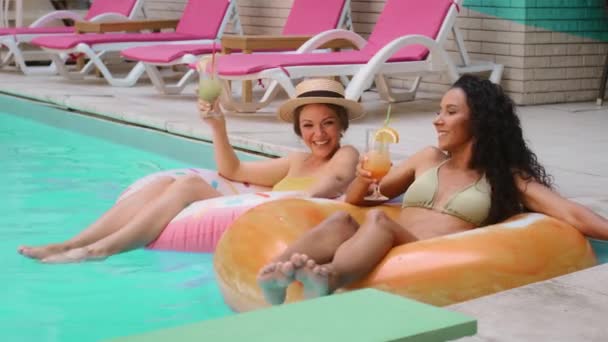 Szczęśliwe śmiejące się kobiety kaukaskie modelki i Arabka świętująca letni weekend w luksusowym hotelu spa przy basenie piją koktajle lodowe. Dwie opalające się dziewczyny pływające na nadmuchiwanych bojach ratunkowych - Materiał filmowy, wideo