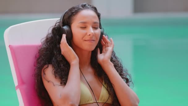 Vyčiněné latina arabština mladá žena student turista krásný dívka modelu sedět na slunci lehátko poslouchat oblíbené hudby ve sluchátkách zpívat píseň relax tanec strávit hudební letní dovolenou v blízkosti bazénu - Záběry, video