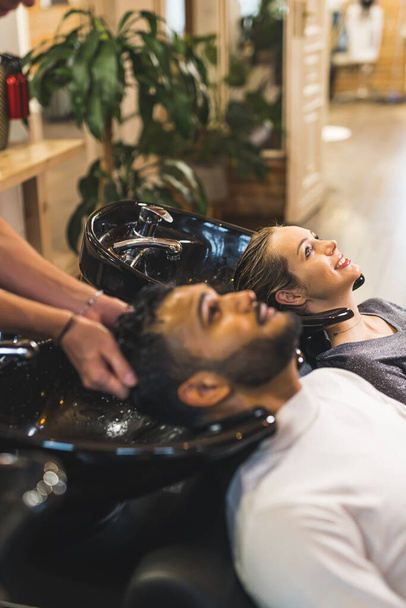 Hintli erkek müşterinin saçı kuaför tarafından profesyonel bir kuaförde sarışın bayan müşterinin yanında yıkanacak. Yüksek kalite fotoğraf  - Fotoğraf, Görsel