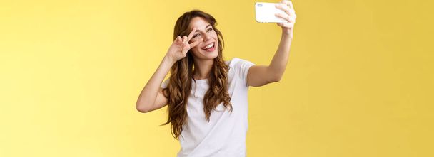 Fröhlich charismatisch gut aussehende gelockte Frau weißes T-Shirt zeigen Friedenssiegerzeichen lächelnd breit posierend zärtlich freundlich Gesichtsausdruck Smartphone-Frontkamera Selfie gelb Hintergrund - Foto, Bild