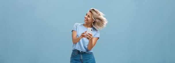 Великие вибрации уносят меня. Беззаботная радостная привлекательная молодая блондинка в стильном наряде машет головой и широко улыбается, слушая музыку в беспроводных наушниках и держа смартфон над синей стеной - Фото, изображение