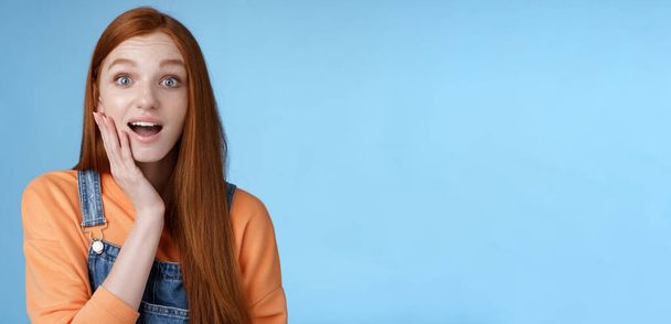 Amazed friendly sensitive impressed redhead girl learn awesome pleasant news open mouth wondered astonished smiling glad friend touch cheek amused, standing blue background happy. Copy space - Photo, Image