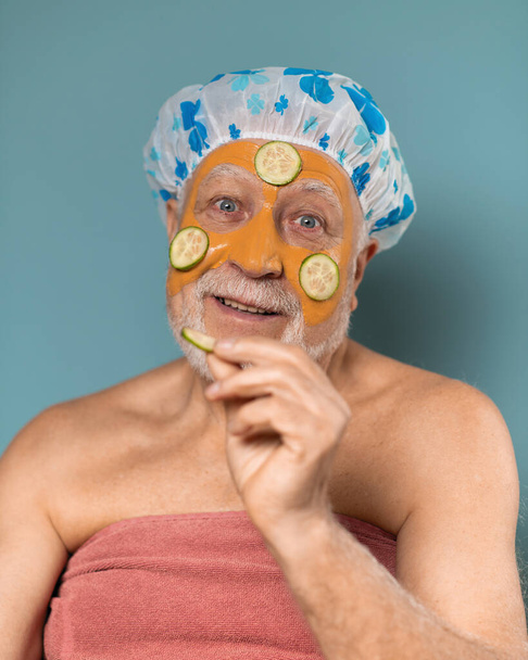 A man with a gray beard aged in a hat for a shower and a towel, with a cosmetic moisturizing facial mask and sliced cucumbers, body care concept, on a blue background, vertical format - Фото, изображение