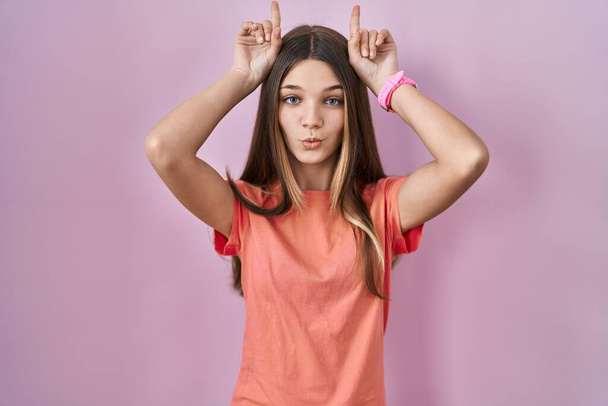Teenager κορίτσι στέκεται πάνω από ροζ φόντο κάνει αστεία χειρονομία με το δάχτυλο πάνω από το κεφάλι ως κέρατα ταύρου  - Φωτογραφία, εικόνα