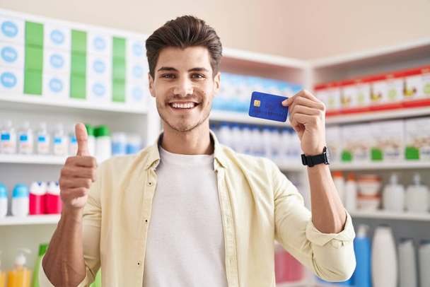 Young hispanic man at pharmacy drugstore holding credit card smiling happy and positive, thumb up doing excellent and approval sign  - Foto, Bild