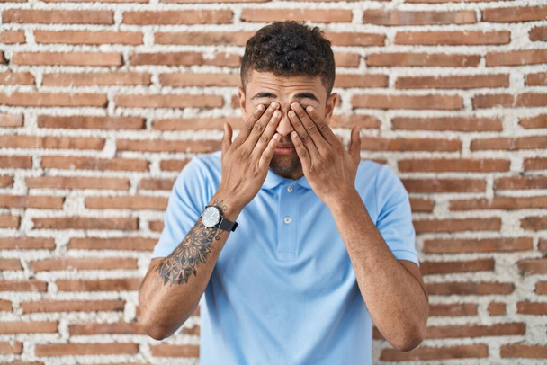 Бразильський молодий чоловік стоїть над цегляною стіною і нищить очі через втому і головний біль, сонний і втомлений вираз. Проблема зору.  - Фото, зображення