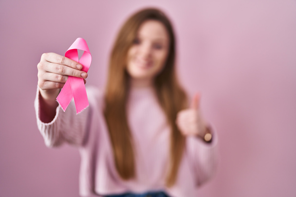 Молодая белая женщина с розовой раковой лентой улыбается счастливой и позитивной, большой палец вверх делает отлично и знак одобрения  - Фото, изображение