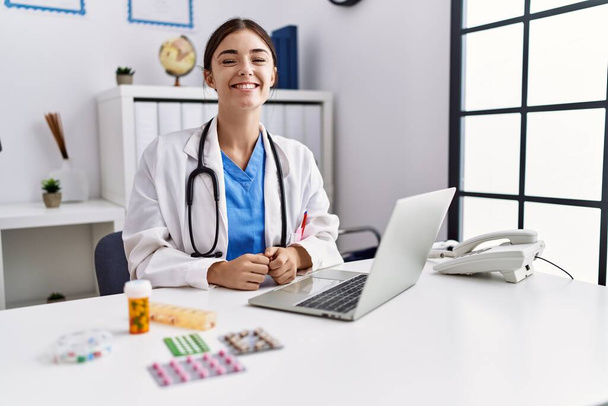 Mladý hispánský lékař žena nosí lékařskou uniformu pracující na klinice vypadá pozitivně a šťastně vstoje a usmívá se s sebevědomým úsměvem ukazující zuby  - Fotografie, Obrázek