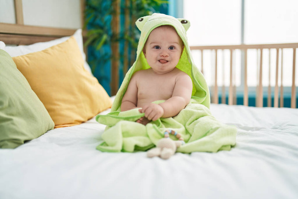 Adorable bebé caucásico con divertida toalla sentada en la cama en el dormitorio - Foto, Imagen