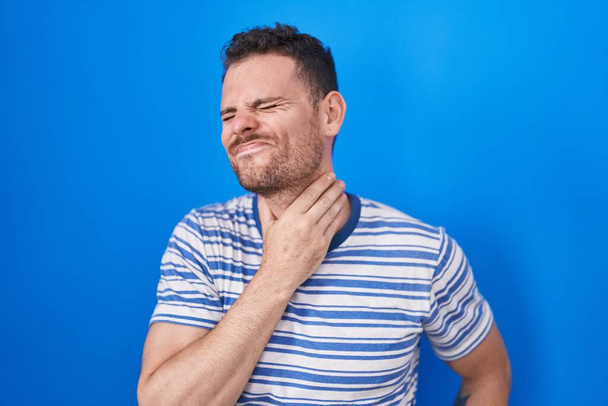Νεαρός Ισπανόφωνος στέκεται πάνω από το μπλε φόντο αγγίζοντας επώδυνο λαιμό, πονόλαιμο για γρίπη, θρόμβο και λοίμωξη  - Φωτογραφία, εικόνα