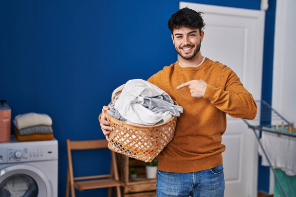 Hispánec s vousy držící koš na prádlo v prádelně s úsměvem ukazující rukou a prstem  - Fotografie, Obrázek