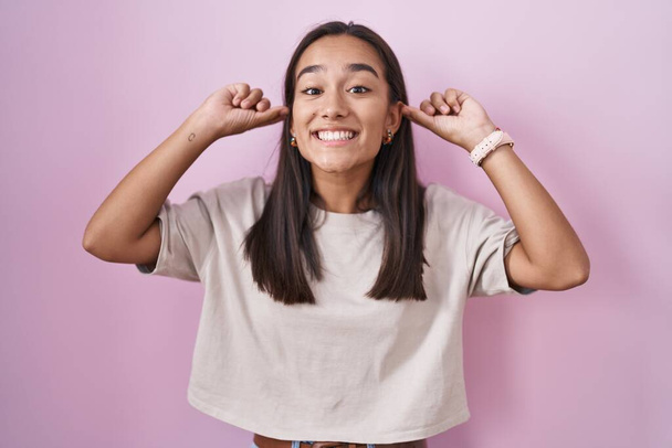 Jonge Spaanse vrouw die met haar vingers over roze achtergrond staat te glimlachen, grappig gebaar. auditieprobleem  - Foto, afbeelding