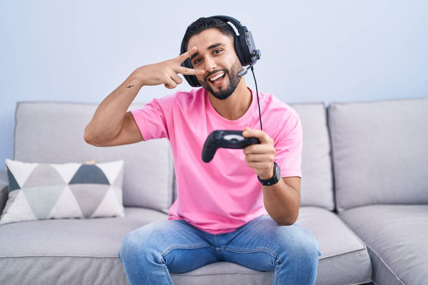 Joven hispano jugando videojuego sosteniendo controlador sentado en el sofá haciendo símbolo de paz con los dedos sobre la cara, sonriendo alegre mostrando la victoria  - Foto, Imagen