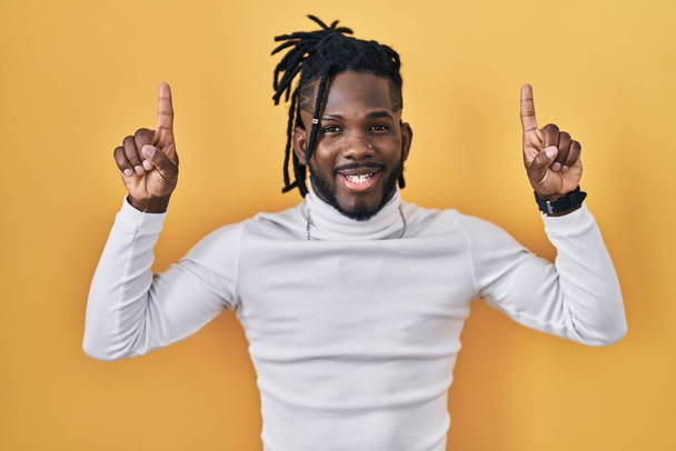 Uomo africano con dreadlocks indossa maglione dolcevita su sfondo giallo sorridente stupito e sorpreso e puntando con le dita e le braccia alzate.  - Foto, immagini