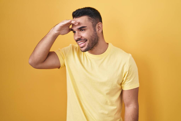 Young hispanic man standing over yellow background very happy and smiling looking far away with hand over head. searching concept.  - Foto, Bild