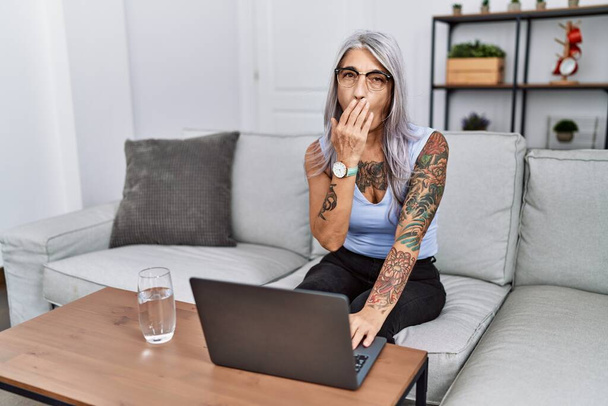 Középkorú, ősz hajú nő, aki laptopot használ otthon, unatkozva ásít, fáradtan takarta el a száját kézzel. nyugtalanság és álmosság.  - Fotó, kép