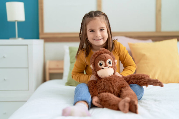 Adorável hispânico menina abraçando macaco boneca sentado na cama no quarto - Foto, Imagem
