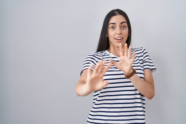 Junge brünette Frau in gestreiftem T-Shirt ängstlich und verängstigt mit Angst Ausdruck Stop-Geste mit den Händen, schreit vor Schreck. Panikkonzept.  - Foto, Bild