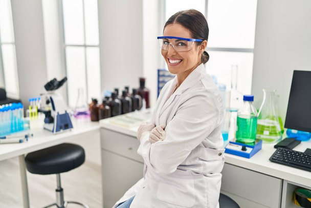 Mladý krásný hispánec žena vědec s úsměvem sedí s rukama zkříženýma gesto v laboratoři - Fotografie, Obrázek