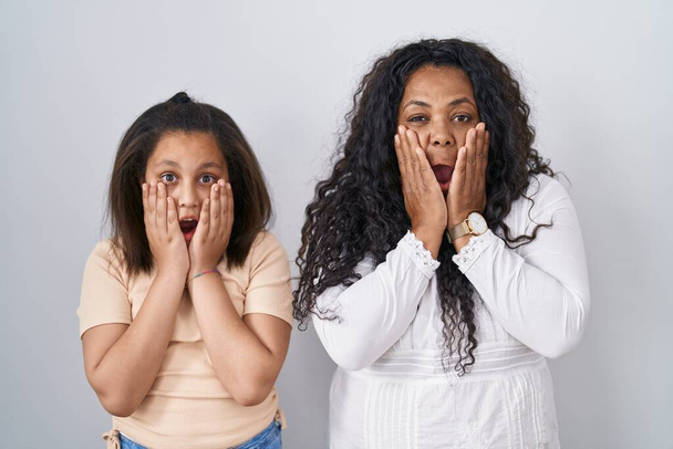 Mother and young daughter standing over white background afraid and shocked, surprise and amazed expression with hands on face  - Фото, зображення