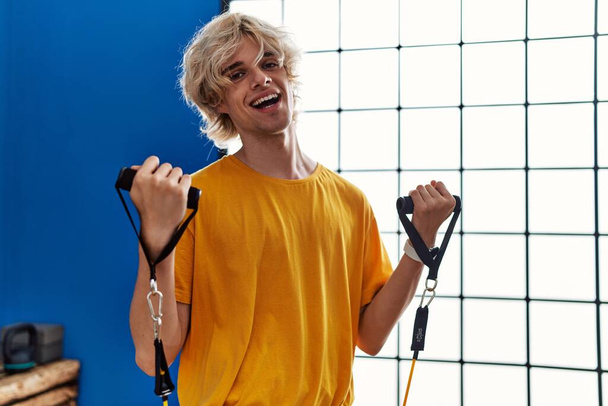 Jeune homme blond souriant confiant en utilisant l'entraînement de bande élastique au centre sportif - Photo, image