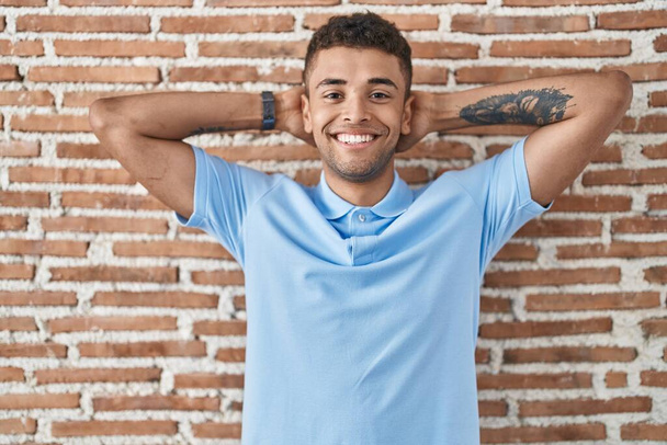 Brazilian young man standing over brick wall relaxing and stretching, arms and hands behind head and neck smiling happy  - Valokuva, kuva