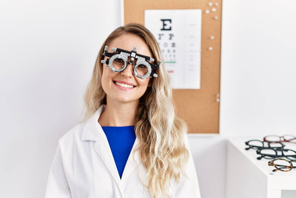 Молодая красивая оптик женщина в очках оптометрии в клинике выглядит позитивно и счастливо стоя и улыбаясь с уверенной улыбкой показывая зубы  - Фото, изображение