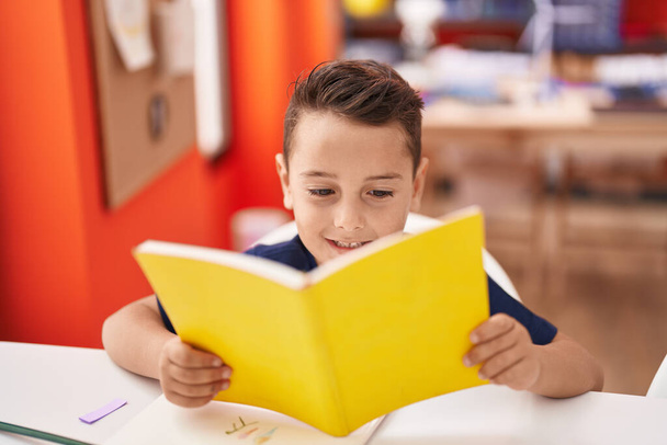 Чарівний іспаномовний малюк студент читає книгу, сидячи на столі в класі
 - Фото, зображення