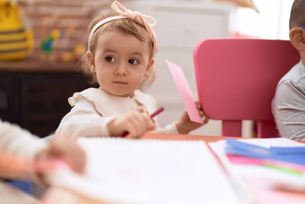 Adorable chica caucásica de pie con expresión relajada sosteniendo papel y lápiz en el jardín de infantes - Foto, Imagen