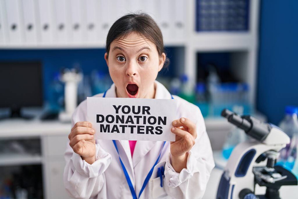 Frau mit Down-Syndrom, die im Labor arbeitet, hält ihr Spendenbanner im Schockgesicht, schaut skeptisch und sarkastisch, überrascht mit offenem Mund  - Foto, Bild