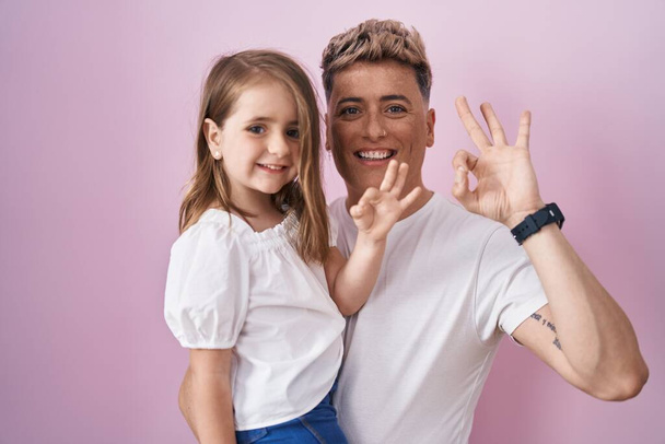Joven padre abrazando hija sobre fondo rosa haciendo signo ok con los dedos, sonriendo gesto amistoso excelente símbolo  - Foto, imagen