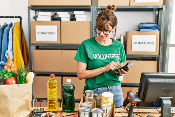 Jonge Spaanse vrouw draagt vrijwilliger uniform met behulp van rekenmachine werken bij het liefdadigheidscentrum - Foto, afbeelding