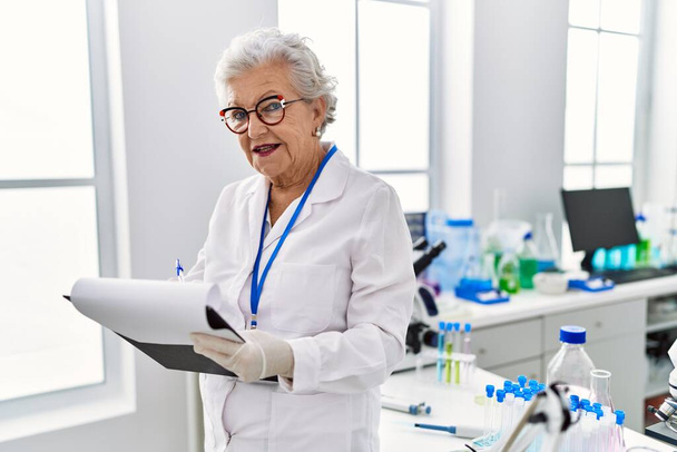 Mujer mayor de cabello gris con uniforme científico escrito en el portapapeles en el laboratorio - Foto, Imagen