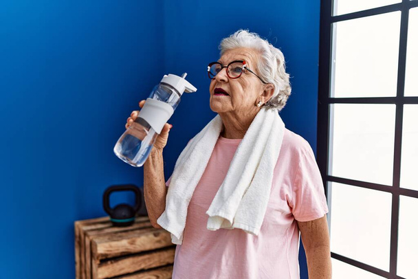 Senior grey-haired woman wearing sportswear holding water bottle at sport center - Photo, Image