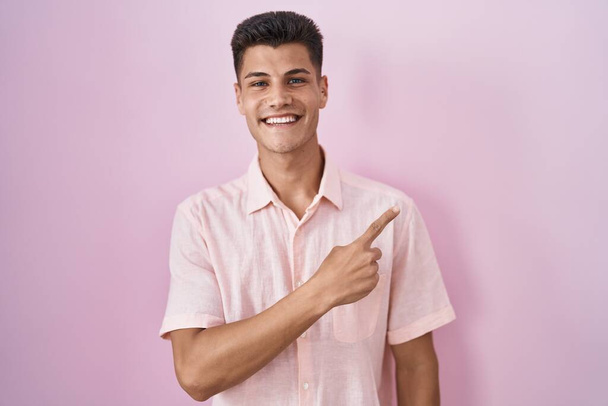Young hispanic man standing over pink background cheerful with a smile on face pointing with hand and finger up to the side with happy and natural expression  - Foto, imagen