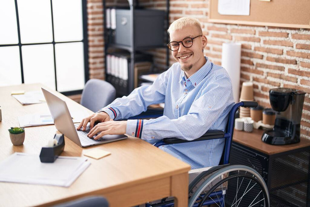 Νεαρός καυκάσιος επιχειρηματίας που κάθεται σε αναπηρική καρέκλα και χρησιμοποιεί φορητό υπολογιστή στο γραφείο - Φωτογραφία, εικόνα