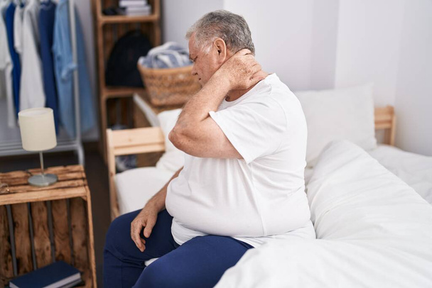 Седой мужчина средних лет, страдающий от травмы шеи, сидит на кровати в спальне - Фото, изображение