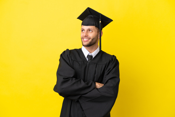 Jeune diplômé universitaire caucasien isolé sur fond jaune levant les yeux tout en souriant - Photo, image