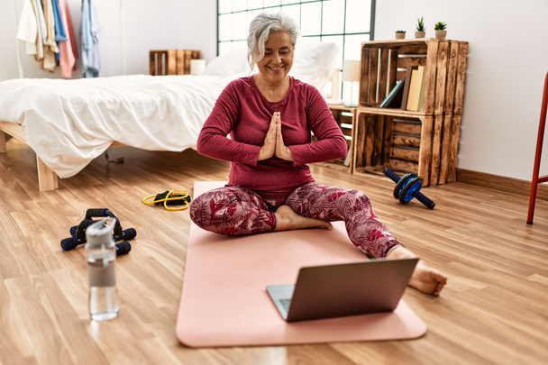 Жінка середнього віку сіроока тренування має відеодзвінок сидить на підлозі в спальні
. - Фото, зображення