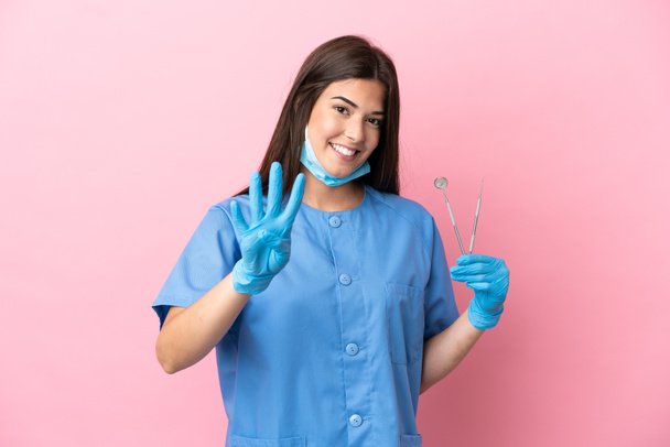 Mujer dentista sosteniendo herramientas aisladas sobre fondo rosa feliz y contando cuatro con los dedos - Foto, imagen