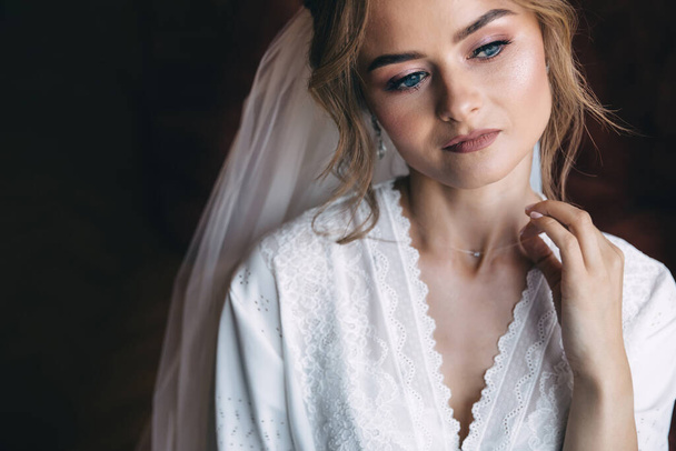 задумчивая невеста в белом халате и вуаль выпрямляет ожерелье вокруг ее шеи. Приготовления утром. - Фото, изображение