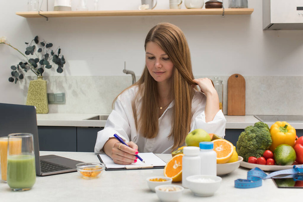 A nutritionist writes an appointment for vitamins and supplements and draws up a nutrition plan during an online consultation with a client, sitting at a table with fresh fruits and vegetables. - Fotoğraf, Görsel