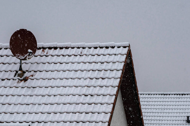 Tiled roof of a house with a satellite dish covered with snow. Gray winter skies and its snowing. - Photo, Image
